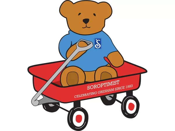 Illustration of a brown teddy bear sitting in a red toy wagon. The bear is wearing a shirt with the Soroptimist logo, a stylized letter S. Text on the wagon reads "Soroptimist / Celebrating Gresham since 1983"