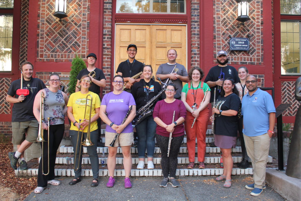 Image of a group of people holding musical instruments standing on the front steps of a brick building.
